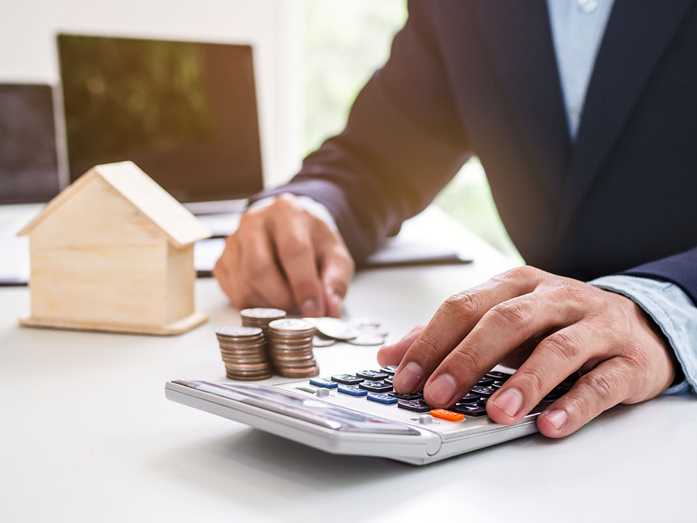 H&auml;nde mit Taschenrechner, M&uuml;nzen und Modellhaus auf dem Schreibtisch zur Immobilienbewertung